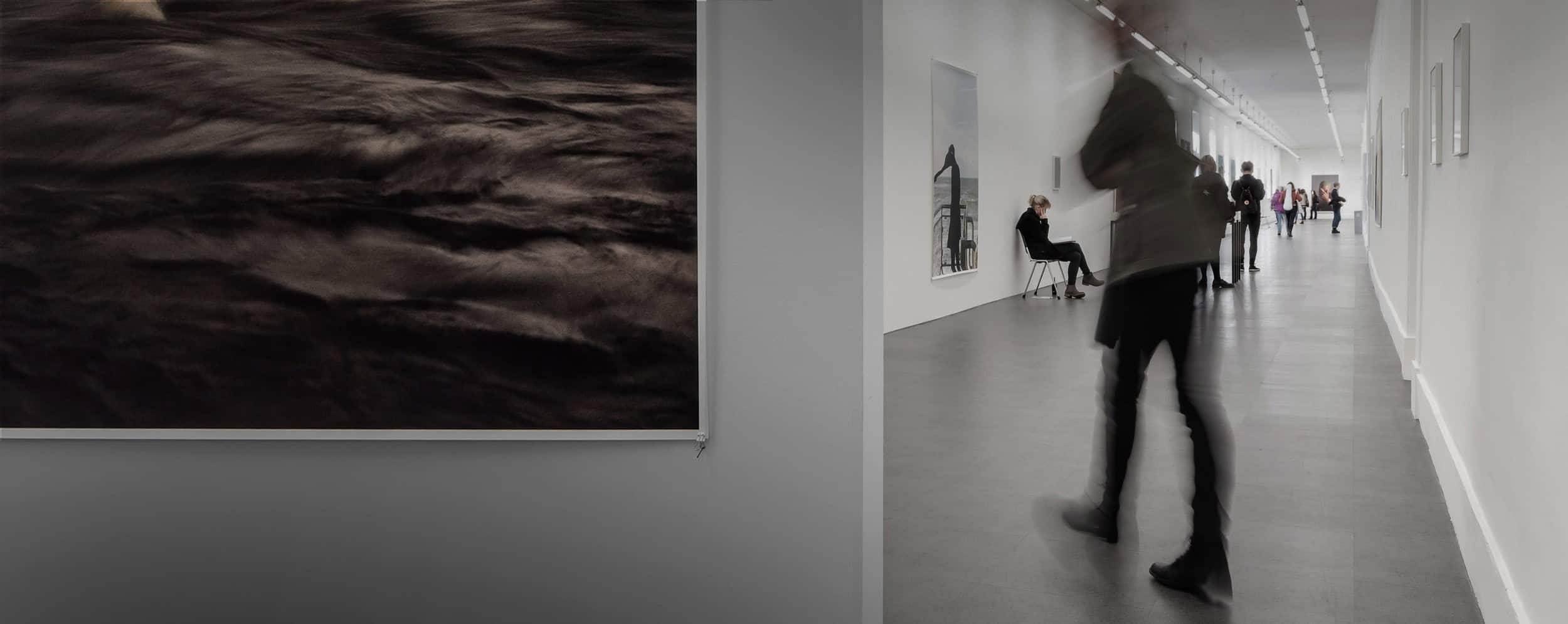 Art gallery scene: Blurred figure walking past a dark, abstract photograph and other art viewers.
