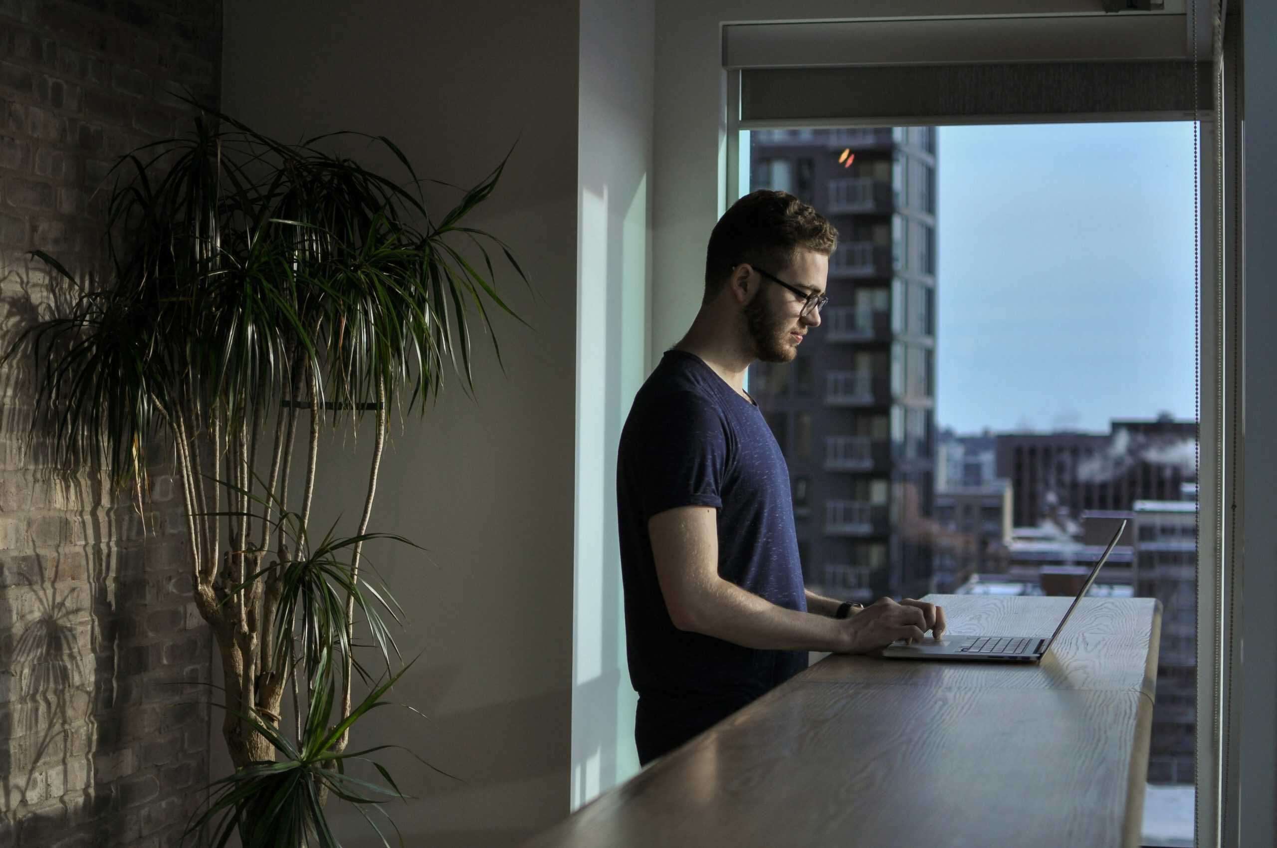 A young man working on a laptop at a high-rise office window, demonstrating modern coworking space, remote work, and professional work environment.